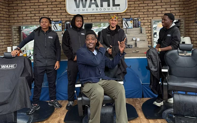 Group of people posing inside a barbershop, with one person seated in a barber chair.