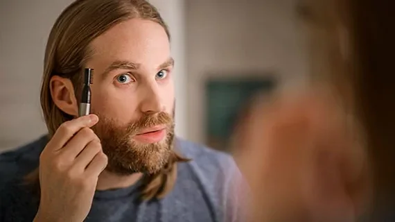 A man looks at his reflection in the mirror as he uses a Wahl trimmer to trim his eyebrows.