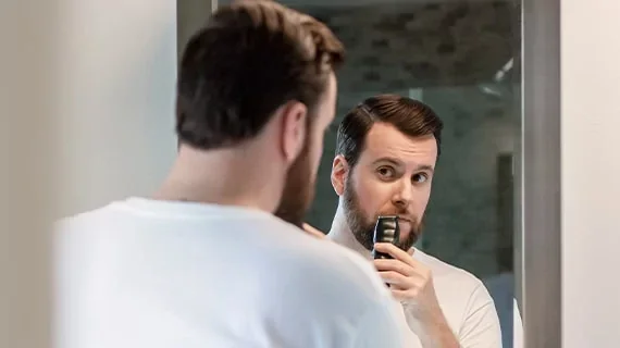 A man looks at his reflection in the mirror as he uses a Wahl trimmer to trim his mustache.