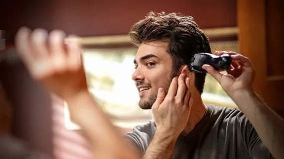 A smiling man uses a Wahl's hair clipper to trim around his ears as he looks into the mirror.