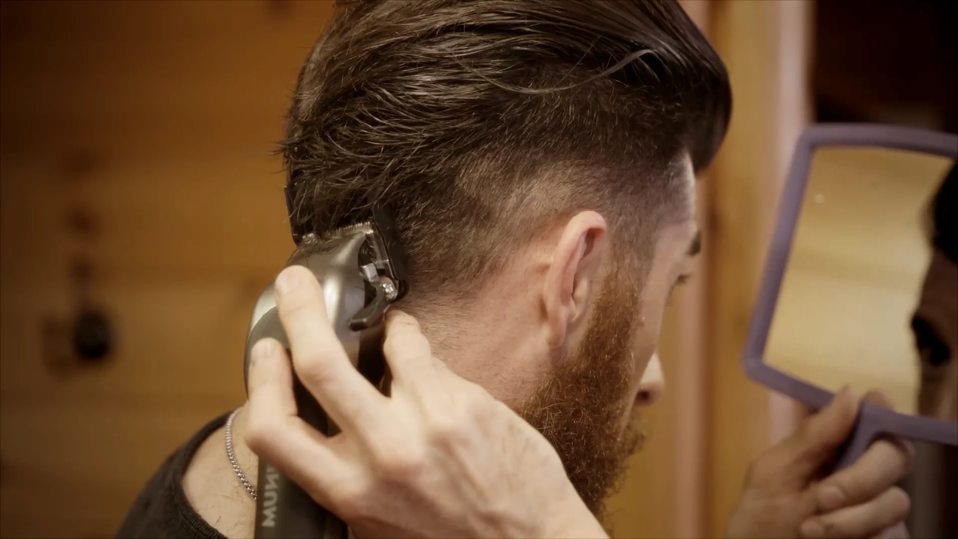 A bearded man using a mirror and Wahl hair clipper to cut the back of his head.
