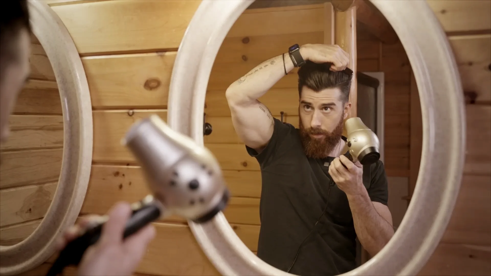 A bearded man using his hands and hair dryer to style his new pompadour haircut.